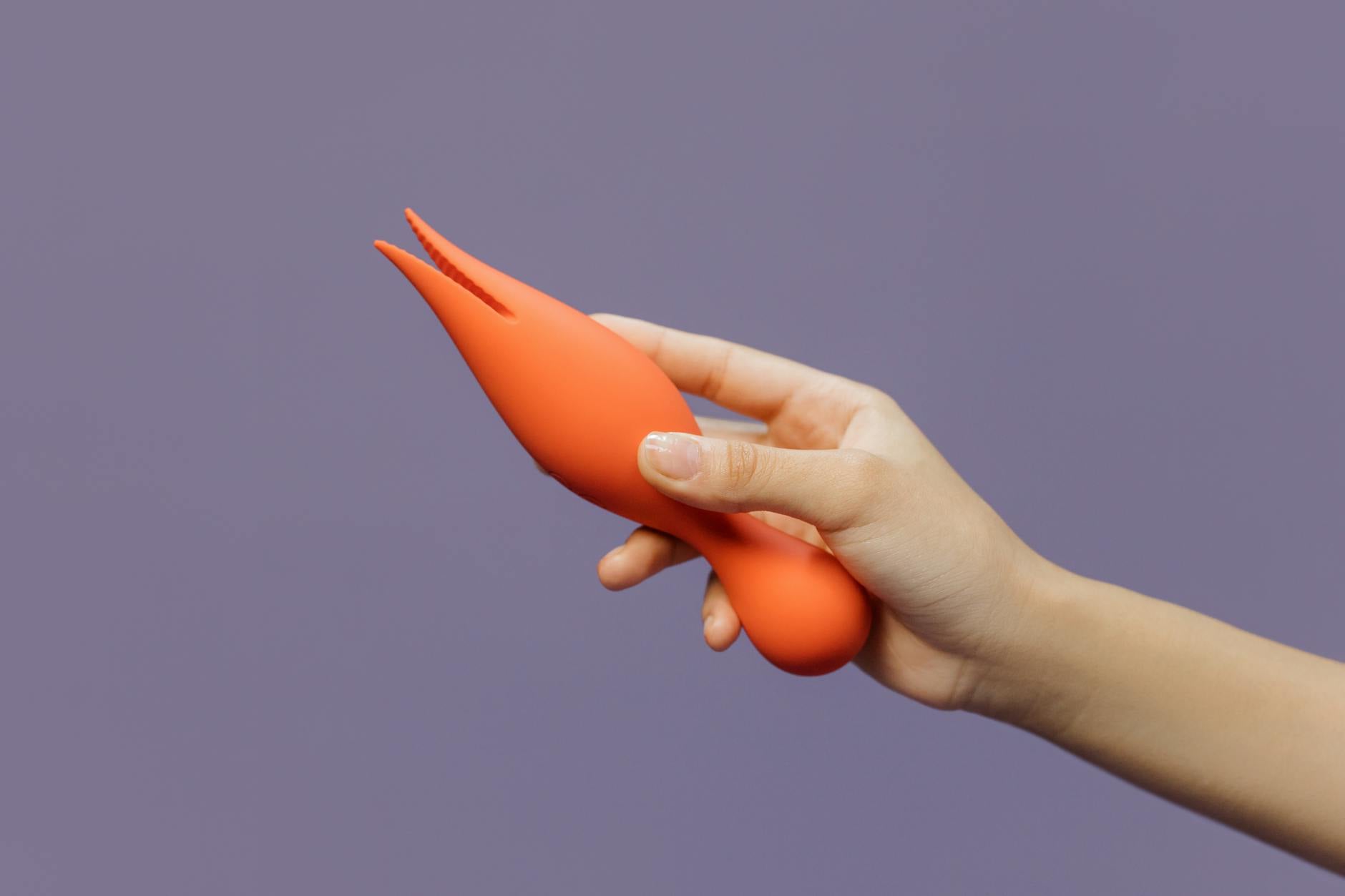 A hand holding a bright orange lemon vibrator against a minimalist purple backdrop, illustrating modern sensual pleasure tools